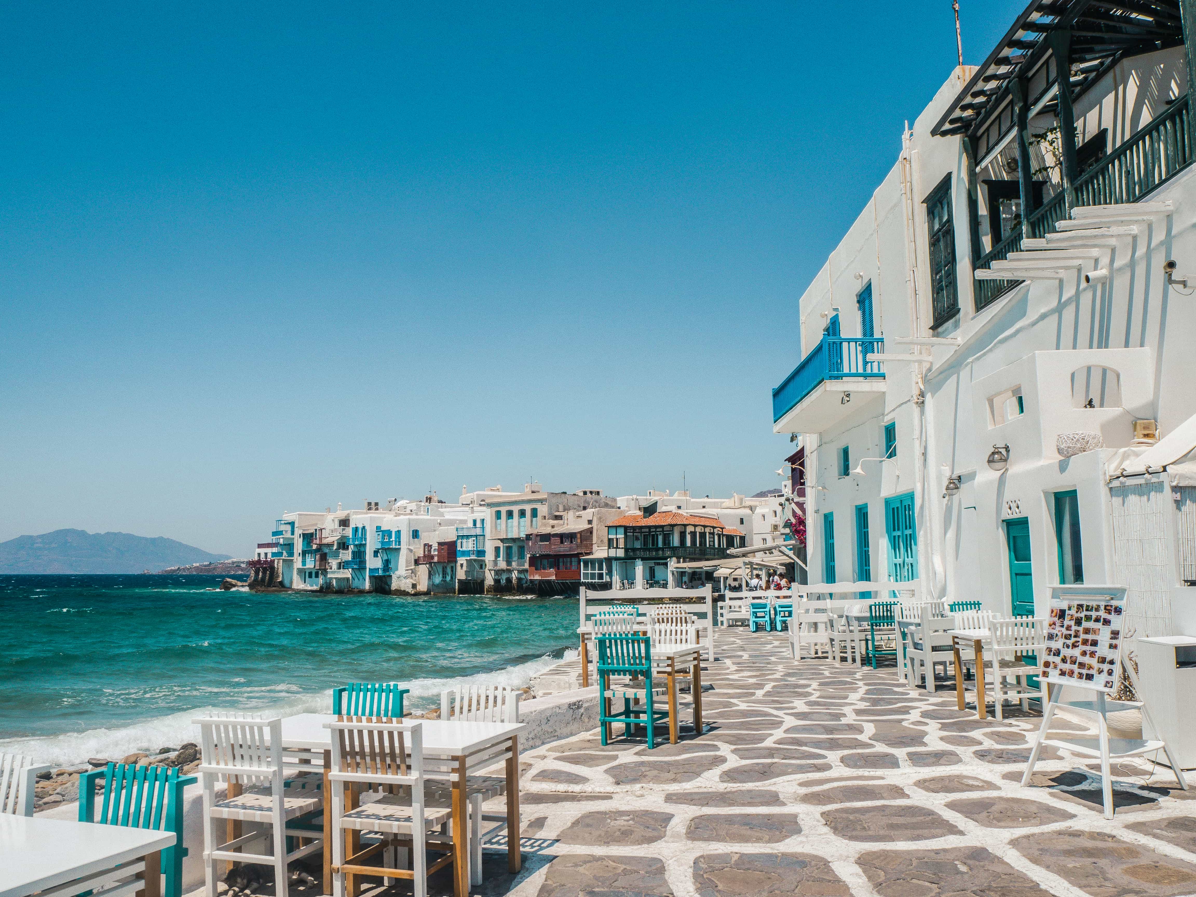 Café tables on the stone waterfront of Little Venice in Mykonos Town, sea spray hitting the quay