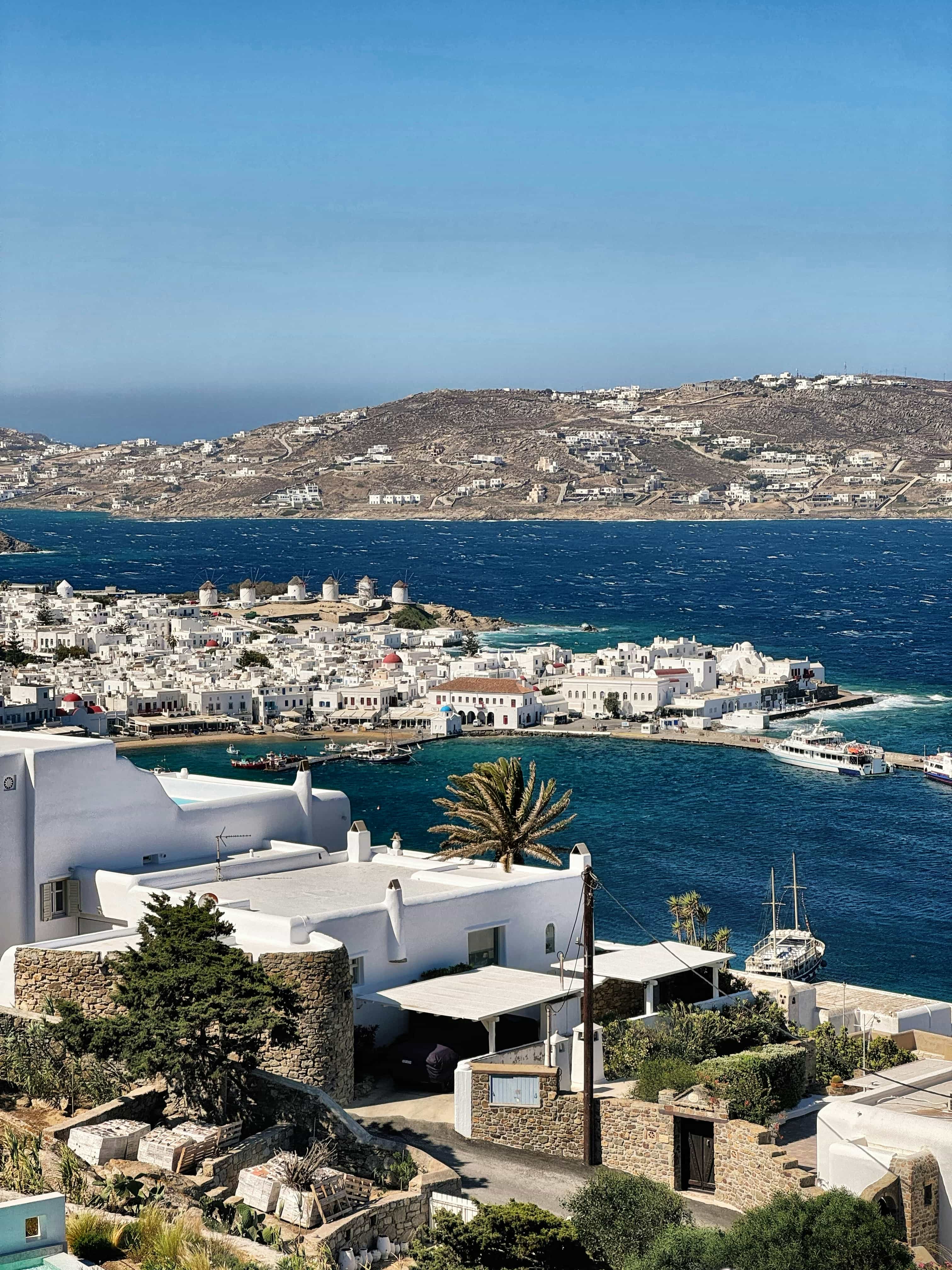 View over the rooftops of Mykonos Town and the old port, with the windmills on the headland